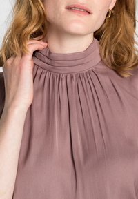 Bledě fialový bezrukávový top s vysokým Kčehem a sklady, vyrobený z jemné tkaniny, která vykazuje měkkou strukturu a decentní lesk.