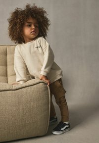 Beige sweatshirt med lös passform, ribbade mudd; cargobyxor i tan med fickor; svarta sneakers med vita sulor; lockigt hår; neutral bakgrund.