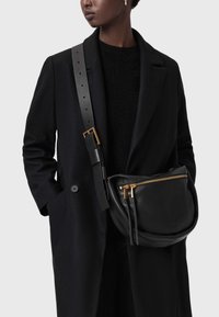 Črna usnjena torbica čez ramo z ukrivljeno obliko, z zlatim zadrgo in dvema potegoma za zadrgo, nošena čez temen plašč z širokim trakom.
