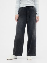 Pantalones de mezclilla negros de pierna ancha con cintura elástica y cordón. Con bolsillos laterales y una textura ligeramente desgastada. Combinados con zapatillas blancas.
