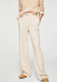 Pantalones de lino de corte ancho en color beige con pliegues delanteros, cintura alta y bolsillos laterales. Combinados con un suéter ligero de punto y zapatos trenzados.