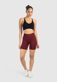 Czarna, krótka sportowa biustonosz z marszczeniami, w zestawieniu z burgundowymi szortami o wysokim stanie. Modelka ma na sobie białe buty sportowe. Gładkie tło.