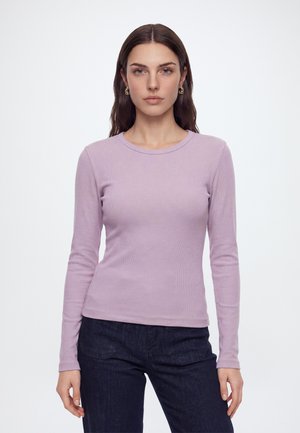 Femme aux longs cheveux bruns portant des boucles d'oreilles dorées, un haut violet clair moulant à manches longues et un jean bleu foncé sur un fond uni.
