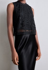 Czarny koronkowy top typu crop z kwiatowymi wzorami, wysokim dekoltem i falbaniastymi brzegami, w połączeniu z gładką czarną satynową spódnicą, tworząc warstwowy wygląd.