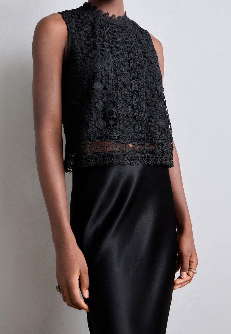 Czarny koronkowy top typu crop z kwiatowymi wzorami, wysokim dekoltem i falbaniastymi brzegami, w połączeniu z gładką czarną satynową spódnicą, tworząc warstwowy wygląd.