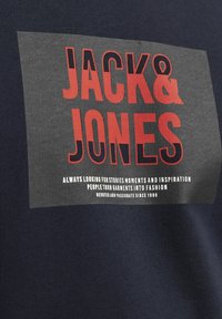 Maritiem kortemouwen T-shirt met een grijze rechthoekige grafiek. Heeft vetgedrukte tekst "JACK & JONES" in rood en wit met extra tekst eronder. Soepele structuur.
