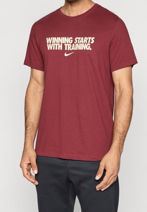 Homme portant un t-shirt Nike bordeaux avec un texte crème "Winning Starts with Training" et un pantalon noir, debout devant un fond uni.