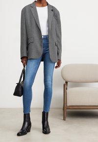 Grå herringbone-blazer över en vit skjorta, tillsammans med blå skinny jeans och svarta klackiga ankelstövlar. Svart handväska i handen.