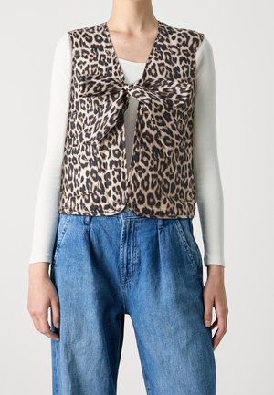 Persoon draagt een mouwloos vest met luipaardprint en een strik aan de voorkant over een wit shirt met lange mouwen en losse blauwe spijkerbroek.