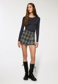 Maglietta a maniche lunghe di colore blu navy abbinata a una gonna avvolgente a quadri grigi, che mostra un design a strati. La modella indossa stivali neri con lacci alti fino al ginocchio.