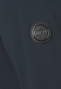 Toppa rotonda nera con logo "COLMAR" su tessuto blu navy testurizzato. Superficie liscia con cuciture visibili intorno al logo.