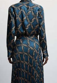 Robe en satin fluide avec un fond bleu orné d'un motif chaîne géométrique beige, manches longues et taille cintrée. Dos plissé.