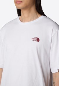 Camiseta de algodón blanca con un pequeño logo burdeos de "The North Face" en el lado izquierdo del pecho. Mangas cortas y cuello redondo.