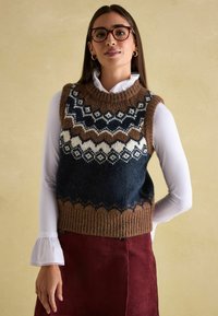 Strickpullover-Weste mit einem geometrischen Muster in dunkelblau, braun und creme. Gerippter Saum und runder Ausschnitt. Über einer weißen Bluse getragen.
