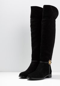 Botas de ante negras, hasta la rodilla, con un acabado texturizado, punta redondeada y un logotipo dorado en el tobillo. Tacón bajo y apilado.