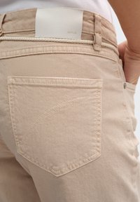 Pantaloni beige con una texture morbida, dotati di una tasca posteriore, cuciture visibili e un'etichetta in pelle bianca impressa con la scritta "OPUS."