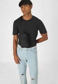 Bolso cruzado negro hecho de tela, con un bolsillo frontal con cremallera, correa ajustable y forma rectangular. Llevado sobre una camiseta negra texturizada y jeans de color azul claro.