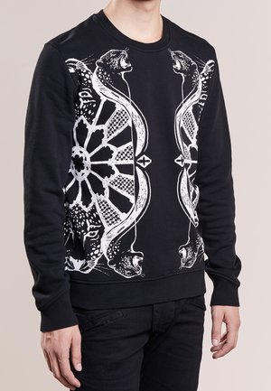 Chemise noire à manches longues avec un motif graphique blanc symétrique de grands félins rugissants et des formes géométriques sur le devant, portée par une personne debout.