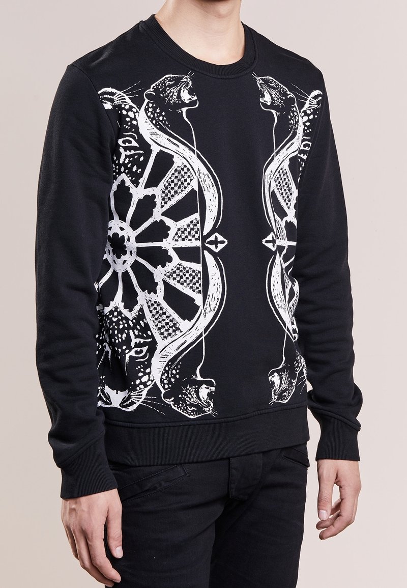 Chemise noire à manches longues avec un motif graphique blanc symétrique de grands félins rugissants et des formes géométriques sur le devant, portée par une personne debout.