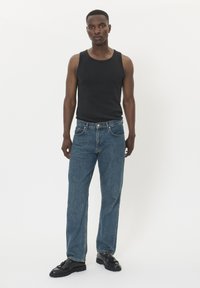 Uomo in piedi contro uno sfondo bianco che indossa una canotta nera, jeans blu e mocassini in pelle nera con nappine.