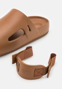 Nike rudos šlepetės be užsegimo, su tekstūruotu paviršiumi, išpjovomis ir nuimamu dirželiu. Dirželis turi logotipą ir audinį kilpelę reguliavimui.