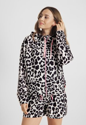 Vrouw die haar haar netjes maakt, gekleed in een zwart-wit gevlekte hoodie en bijpassende korte broek met roze rits en trekkoorden tegen een effen achtergrond.