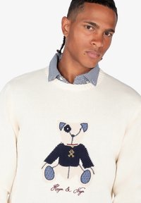 Suéter blanco de punto con un diseño de oso bordado, camisa azul marino, acentos azules y costura detallada. Botones en el collar de la camisa.