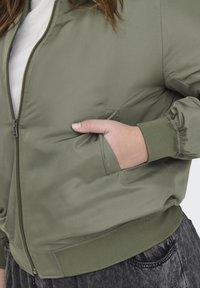 Olivovo zelená bomber bunda so zipsovým uzatváraním, rebrovanými manžetami a pásom. Obsahuje bočné vrecká a jemnú, ľahkú textúru.