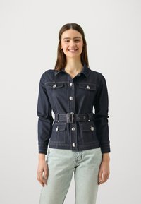 Mørk denimjakke med knappelukking, lange ermer og to brystlommer. Har belte i midjen og kontrastsøm detaljer.