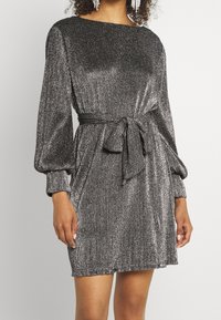 Silver shimmer-klänning med båtformad halsringning, långa puffärmar och en midjebanddetalj. Tyget har en texturerad metallisk yta.