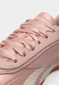 Κοντινή λήψη ενός ροζ αθλητικού παπουτσιού Reebok με σατέν κορδόνια και υφή από σουέντ, που δείχνει το λογότυπο κοντά στη μαλακή γλώσσα και το λευκό χείλος της σόλας.