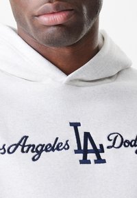 Hoodie cinza claro feito de um tecido suave. Apresenta texto em azul escuro "Los Angeles Dodgers" e o logotipo "LA", com um bolso tipo canguru na frente.