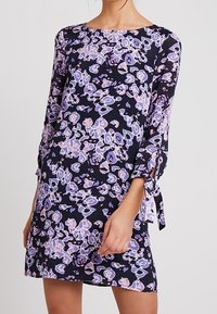 Kobieta prezentuje granatową sukienkę ozdobioną fioletowymi i białymi wzorami kwiatowymi, z długimi rękawami zakończonymi wiązaniami przy mankietach.