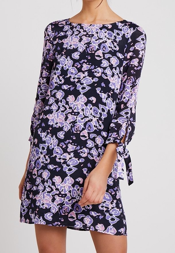 Kobieta prezentuje granatową sukienkę ozdobioną fioletowymi i białymi wzorami kwiatowymi, z długimi rękawami zakończonymi wiązaniami przy mankietach.