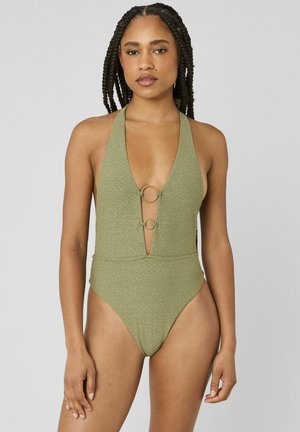 Femme aux cheveux tressés portant un maillot de bain licou texturé vert olive avec deux anneaux décoratifs dorés sur le devant, debout devant un fond uni.
