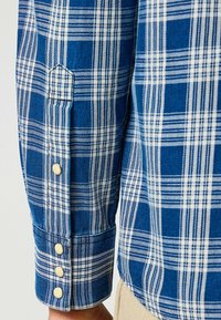 Camisa de cuadros azul y blanca con puño abotonado y tela texturizada. Costuras visibles y detalle de manga doblada.