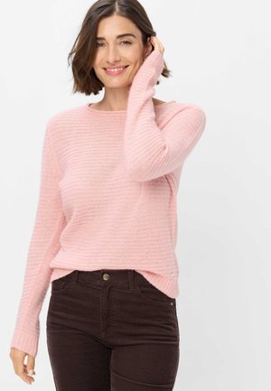Donna con capelli castani corti sorridente, indossa un maglione lavorato a maglia rosa chiaro e pantaloni di velluto a coste marrone scuro su sfondo bianco.