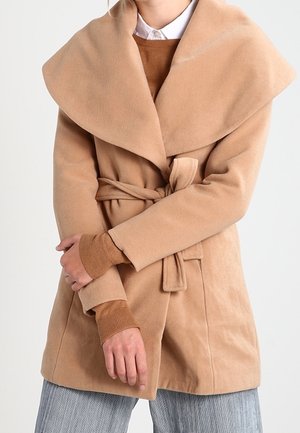 Kvinde iført en kamelfarvet kåbe med bælte over en brun sweater og hvid skjorte med krave, med grå bukser synlige nedenunder.