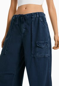 Laivastonsiniset cargo-housut, joissa on joustava vyötärö, kiristysnauha, sivutaskut ja läpätasku oikeassa lahkeessa. Valmistettu kestävästä kankaasta.