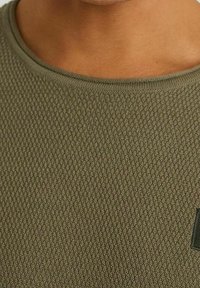 Sweter w oliwkowym zielonym kolorze o fakturze, z okrągłym dekoltem, posiadający wzór w wafle i mały detal z logo. Gładkie krawędzie i miękki materiał.