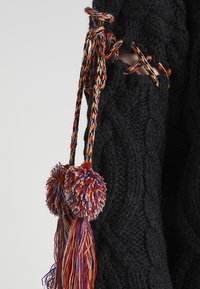 Maglione nero lavorato a maglia con un motivo a treccia, dotato di colorati lacci intrecciati e due grandi pompon nei toni di rosso, arancione e blu.