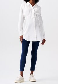 Femme enceinte portant une chemise de maternité blanche boutonnée, des leggings navy et des baskets blanches, se tenant devant un fond uni.