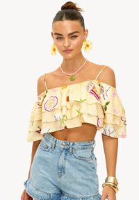 Haut court jaune épaule dénudée avec des couches à volants, orné d'imprimés colorés de fruits et d'étoiles de mer. Associé à un short en denim taille haute.