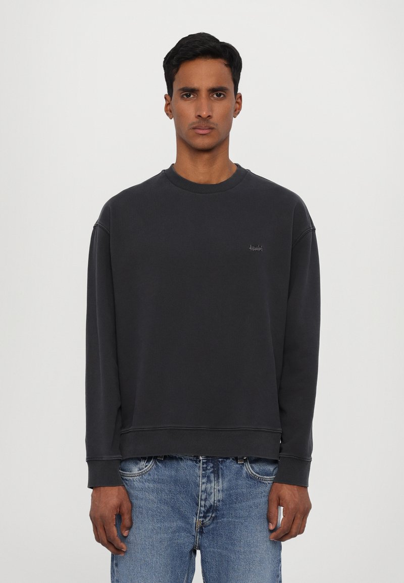 Sweatshirt gris foncé avec un col rond, des poignets et un ourlet côtelés. Présente un petit logo sur la poitrine. Porté avec un jean bleu clair.