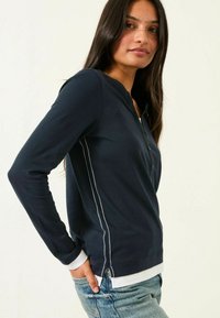 Marineblaues Langarmoberteil mit einem Henley-Ausschnitt, kontrastierenden weißen Nähten an den Seiten und einem weißen Saum am unteren Ende.