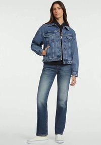 Kvinna stående i en blå denimjacka, svart huvtröja, blå jeans och vita sneakers mot en enfärgad vit bakgrund.