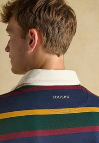 Gestreiftes Polohemd mit navy, grün, kastanienbraun und gelben Linien, mit einem weißen Kragen. "JOULES" ist auf dem Rücken gestickt.