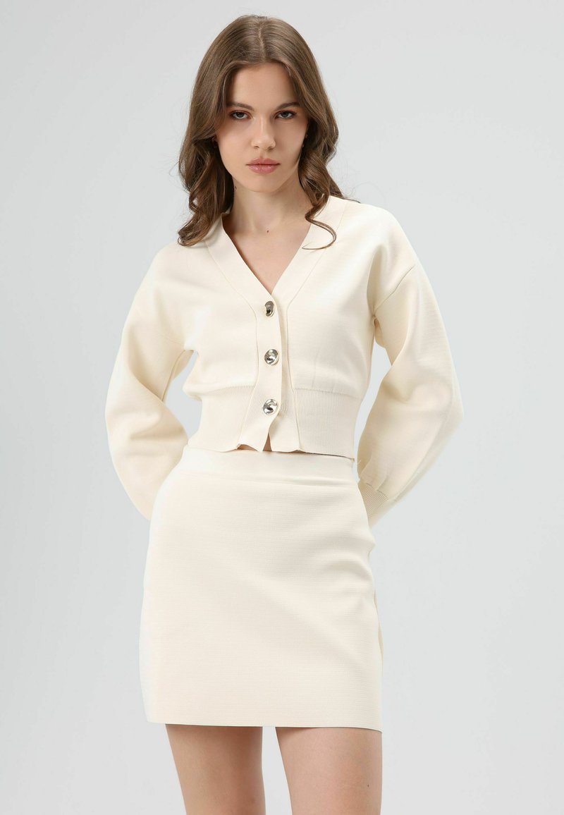 Roomkleurig cropped vest met V-hals, vijf zilveren knopen en lange mouwen, gecombineerd met een bijpassende minirok, beide van een gestructureerde stof.