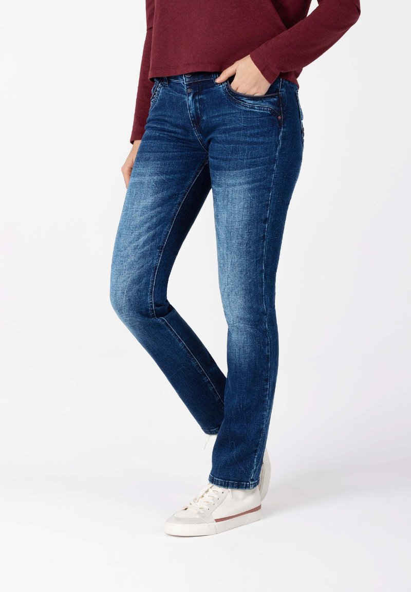 Dunkelblaue Jeans im Slim-Fit-Stil mit fünf Taschen, sichtbaren Nähten und einer leichten Ausbleichung an den Oberschenkeln. Kombiniert mit weißen Sneakers.
