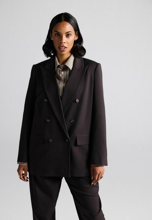 Dvojřadový černý blazer vyrobený z texturované látky se dvěma předními kapsami, zkombinovaný s pruhovanou košilí na knoflíky pod ním.